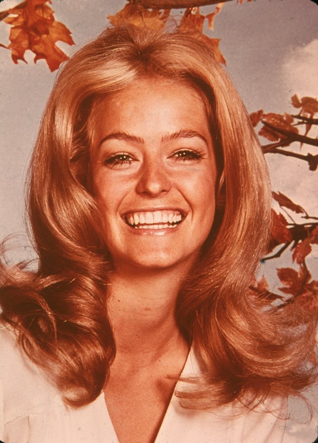 tendencia-sobrancelha-anos-70 Foto da atriz Farrah Fawcett nos anos 70. Ela está com uma camiseta branca, cabelo solto e maquiagem leve. Ela olha para a câmera e sorri.