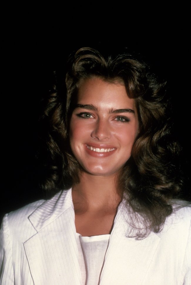 tendencia-sobrancelha-anos-80 Brooke Shields nos anos 80. Ela usa uma camiseta e casaco branco, cabelo solto e maquiagem natural. Ela olha para a câmera e sorri.
