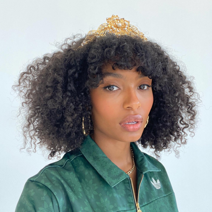 Yara Shahidi com expressão facial surpresa usando franja e uma jaqueta verde e uma coroa dourada na cabeça