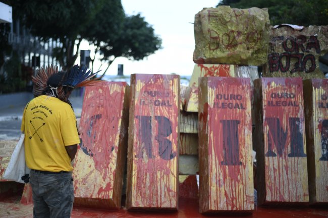 Protesto de povos indígenas contra o garimpo ilegal. Eles seguram cartazes e a bandeira do Brasil cobertos de tinta vermelha, que representa o sangue