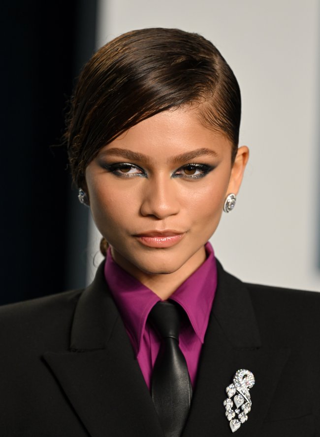 zendaya-na-vanity-fair-com-look-com-gravata Foto da Zendaya na Vanity Fair de 2022 com um look com blazer e gravata e maquiagem com esfumado preto nos olhos. Ela olha para o lado e não sorri.