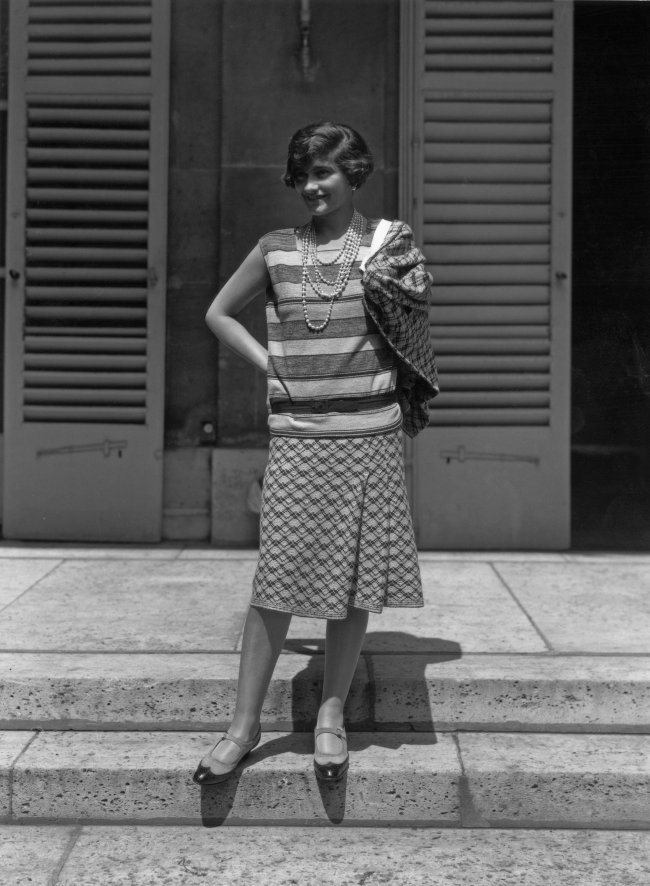 coco-chanel-sapatos-mary-jane Foto em preto e branco da Coco Chanel de 1929. Ela usa uma camisa larguinha de listras, saia midi estampada e sapatos mary jane. Ela olha para o lado e sorri levemente.