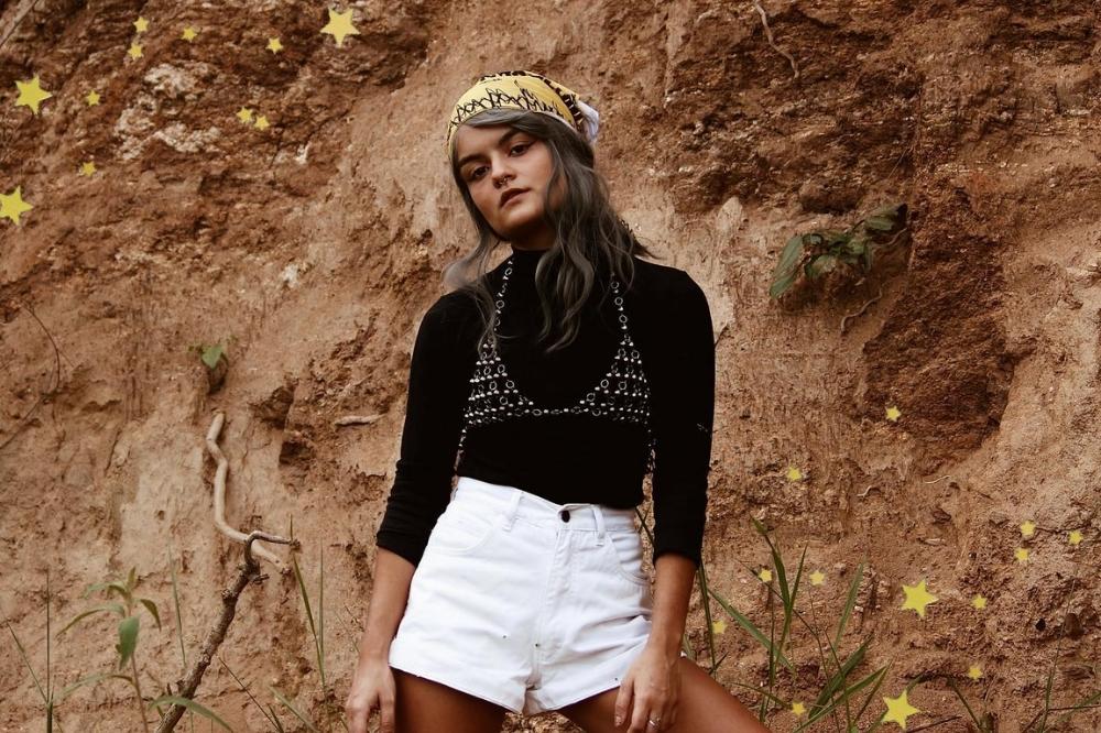 Foto de uma mulher com a cabeça levemente inclinada para a direita que não sorri para a foto. Ela usa uma blusa de manga longa preta, peça de metal como sobreposição, short branco e lenço amarrado na cabeça.
