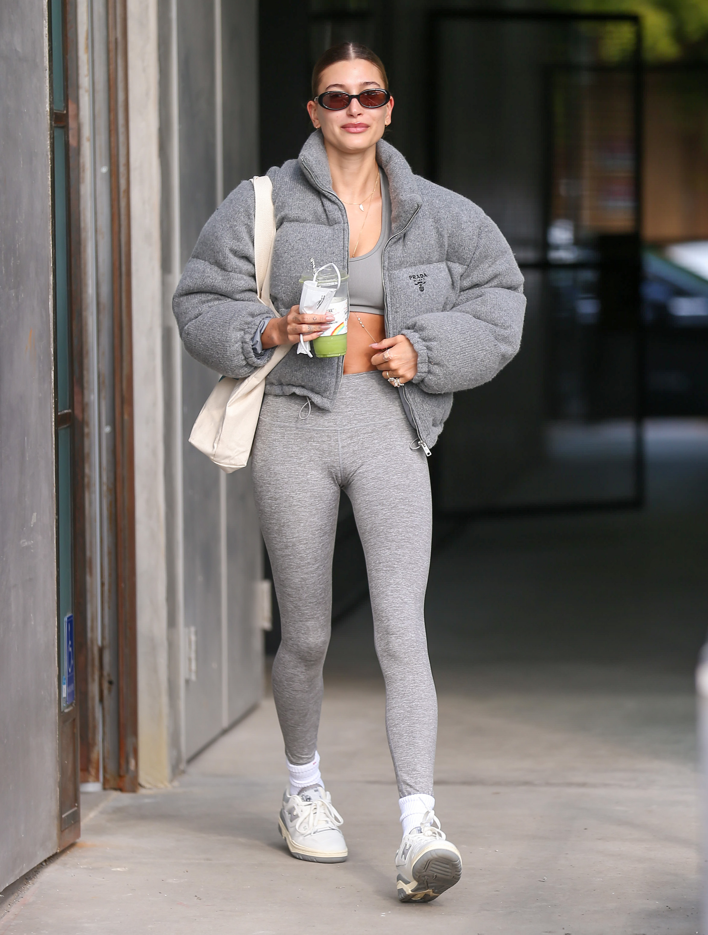 Celebrity Sightings In Los Angeles – January 08, 2022 Hailey Bieber usando look todo cinza com jaqueta puffer, top, legging, tênis branco, com uma ecobag em um dos ombros e um óculos de sol