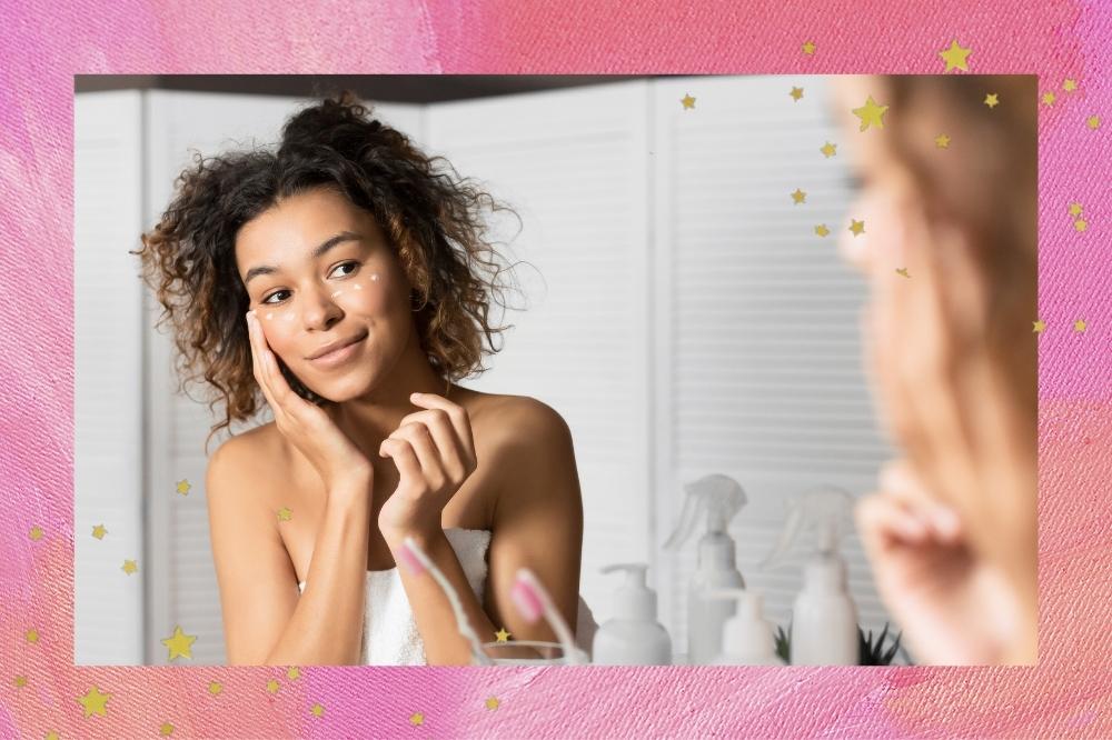 Montagem com o fundo rosa e detalhe de estrelas douradas nas bordas com a foto de uma mulher de frente para o espelho. Ela está com uma toalha branca, cabelo solto, pele limpa e aplica um creme com a mão na região dos olhos. Ela olha para o espelho e sorri levemente.