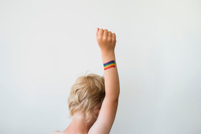Medo crônico: saúde mental da população LGBTQIA+ precisa de atenção extra Pessoa branca de cabelos loiros de costas, com o braço direito levantado e um arco-íris pintado na mão