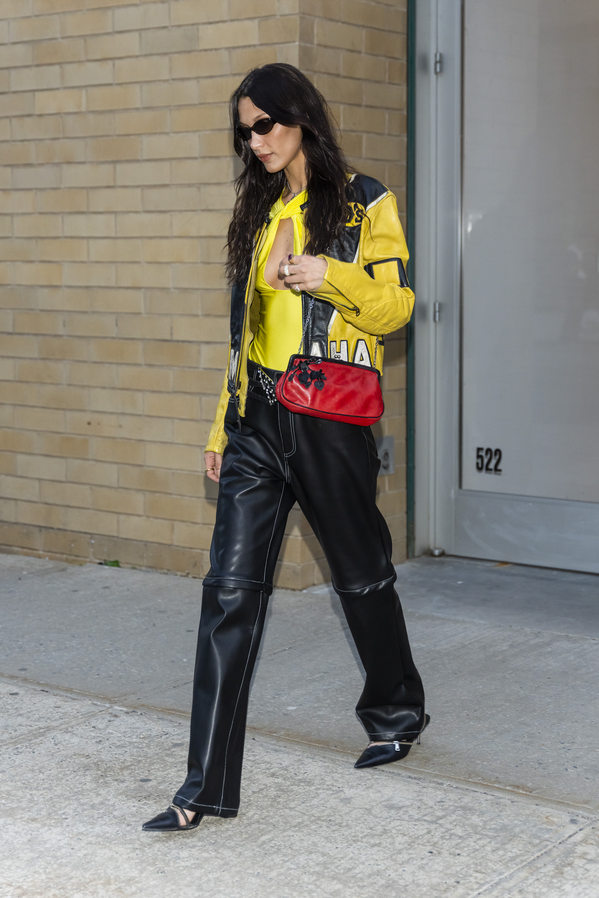 Celebrity Sightings In New York City – May 10, 2022 Bella Hadid usando look com referências da estética bikercore com jaqueta de couro amarela e preta, calça de couro e bolsa vermelha