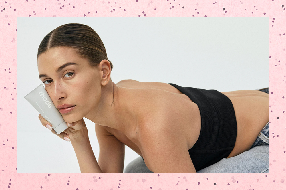 Montagem em fundo rosa de foto de Hailey Bieber deitada segurando um produto da sua marca de pele Rhode e usando um top faixa preto