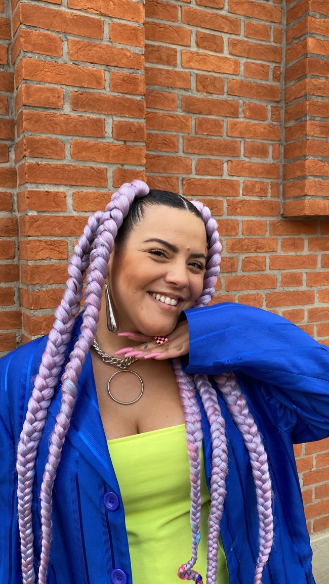 look-colorido-tranças-e-maquiagem-de-gab-alves-spfw Imagem mostra Gab Alves sorrindo com a mão direita no queixo enquanto usa tranças e look colorido na spfw