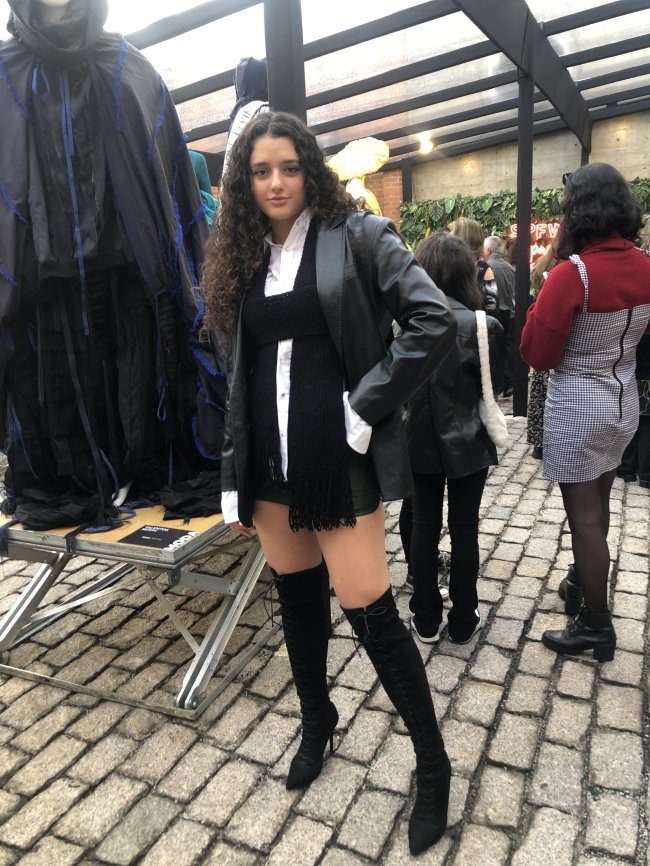 look-com-couro-spfwn53 Foto da Giovanna Venturoli na São Paulo Fashion Week nº53 usando blazer de couro, camisa branca, saia preta e bota preta over the knee. Ela olha para a câmera e sorri.