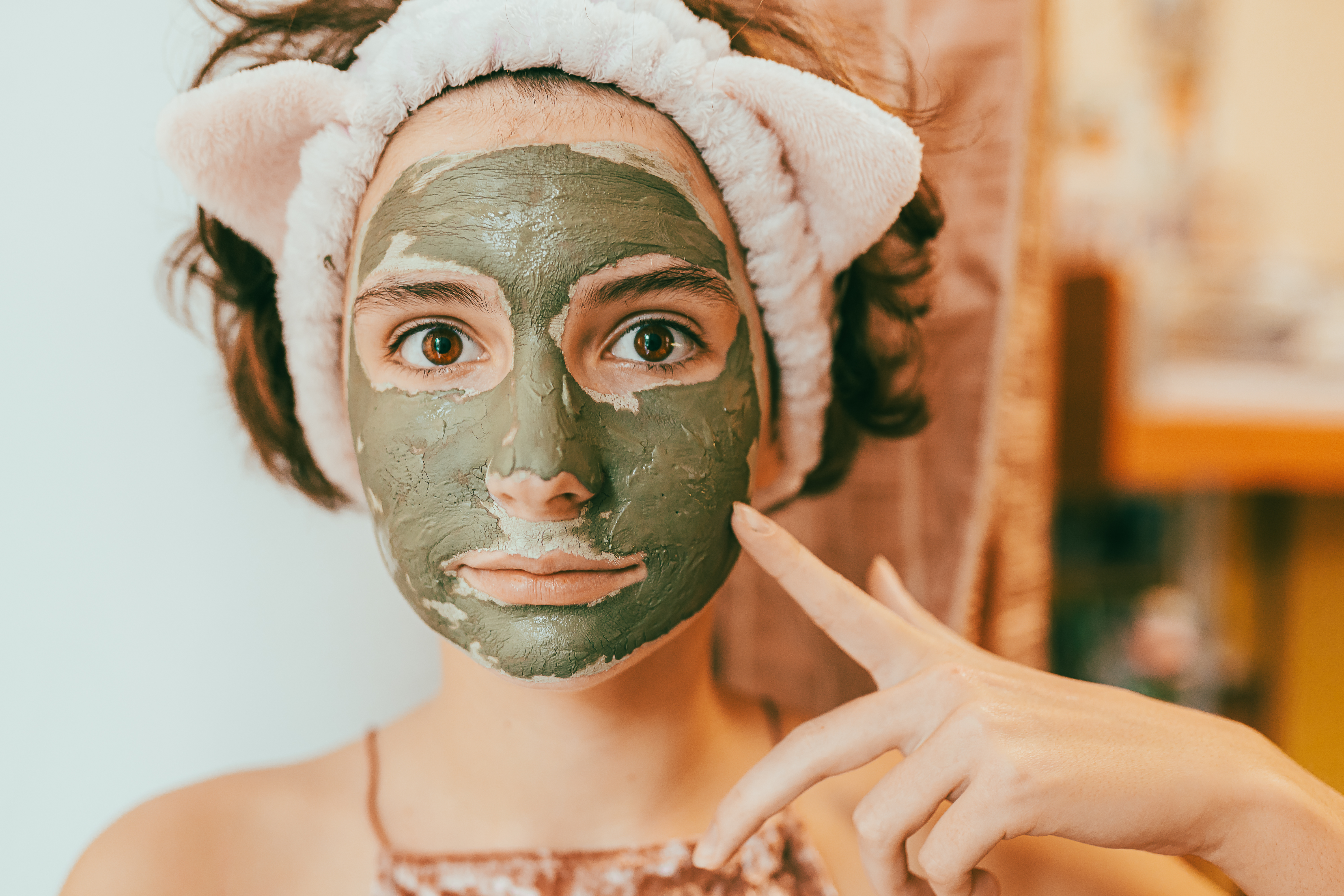 Application of cosmetic procedures for teenage skin. Face of girl close-up with applied mask Garota com máscara facial de argila verde, expressão facial séria e uma faixa no cabelo