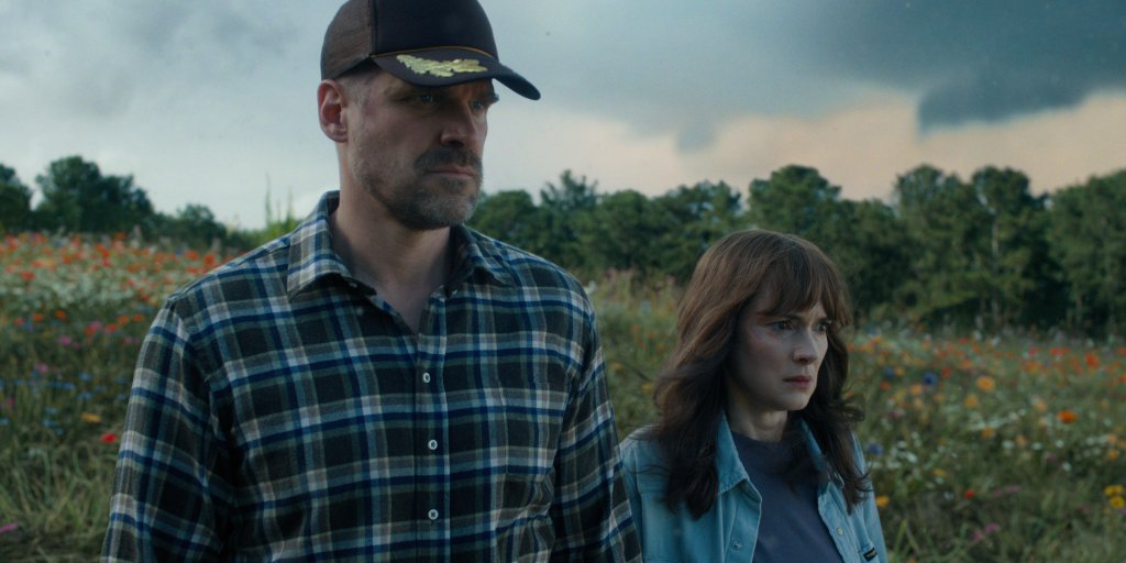 David Harbour como Jim Hopper e Winona Ryder como Joyce Byers em Stranger Things; eles estão com expressões sérias olhando para frente em uma área aberta e verde