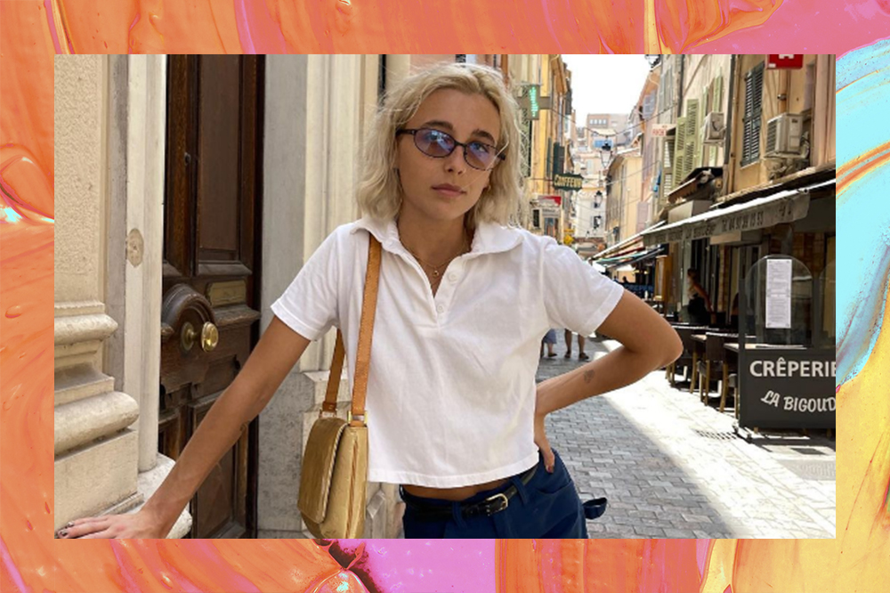 Foto de Emma Chamberlain usando camiseta polo branca, short jeans, óculos de sol, bolsa pendurada no ombro e uma mão na cintura. O fundo da montagem é laranja, amarelo, rosa e azul
