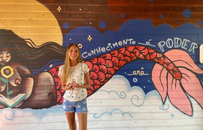 No Amazonas, estudantes ribeirinhos vão até a escola para ter aulas EAD Estudante ribeirinha do Amazonas posa em frente a um mural com os dizeres
