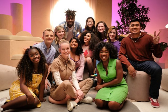 Grupo de pessoas posando para foto sorrindo enquanto estão sentados com uma luz forte em formato de círculo ao fundo e luzes rosas nas laterais