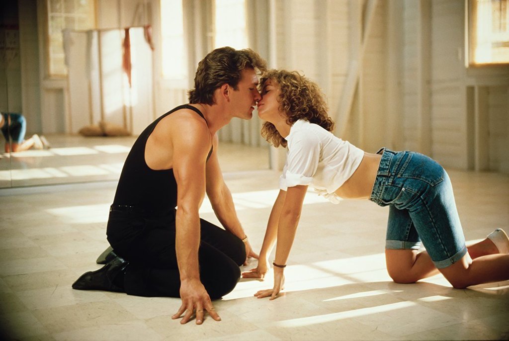 Casal do filme Dirty Dancing no chão, ajoelhados, dando um selinho; eles vestem roupas de ginástica