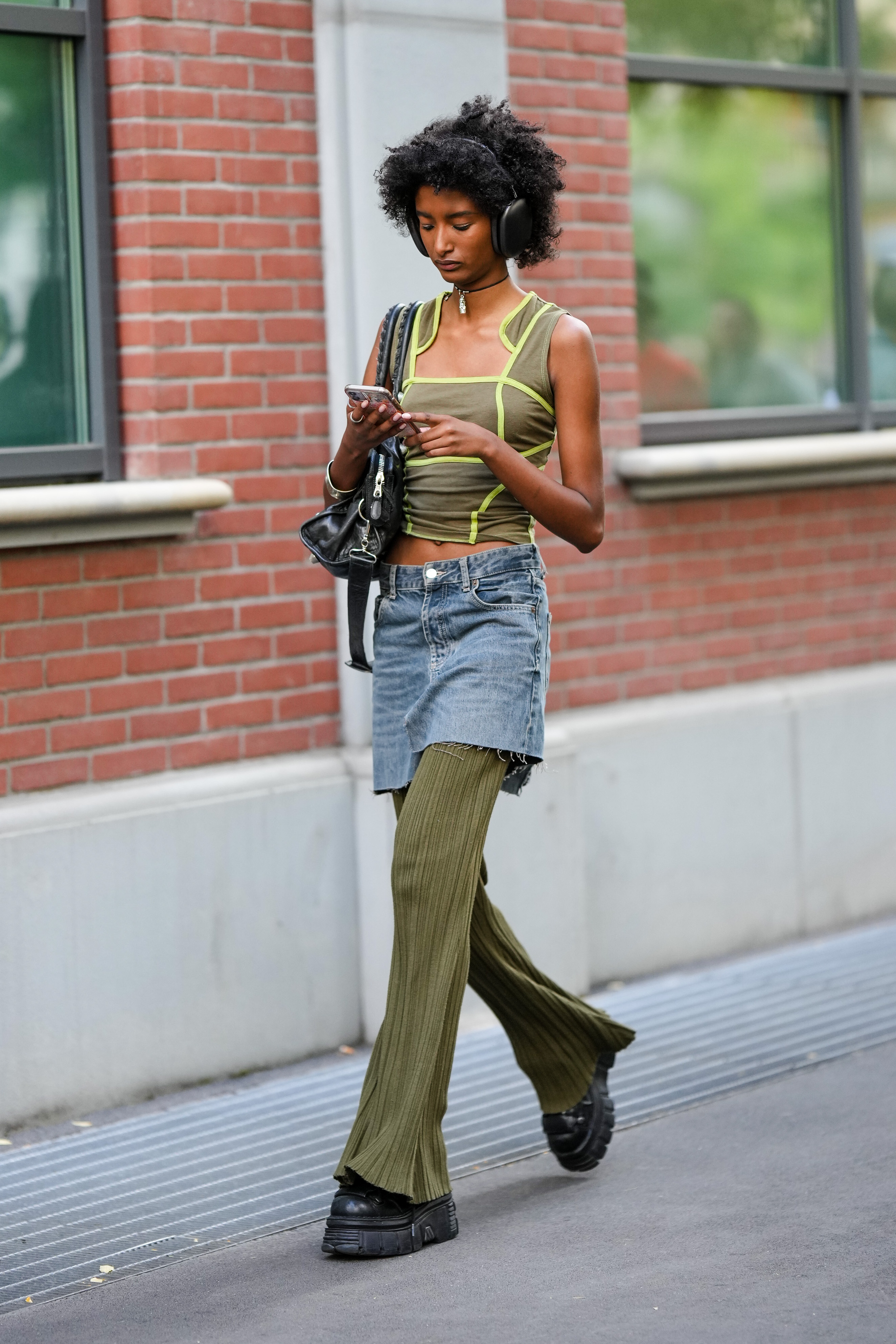 StreetStyle – Day 2 – Milan Fashion Week Womenswear Spring/Summer 2023 Garota usando top verde, saia jeans por cima de calça flare verde, tênis preto, bolsa preta pendurada em um dos ombros e headphone preto enquanto olha para o celular e anda na rua na semana de moda de Milão