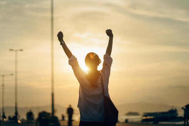 10 dicas de passeios para você fazer sozinha e curtir a própria companhia Uma mulher celebra a vida com as mãos erguidas para o alto. Ela está de costas e ocorre um pôr-do-Sol ao fundo