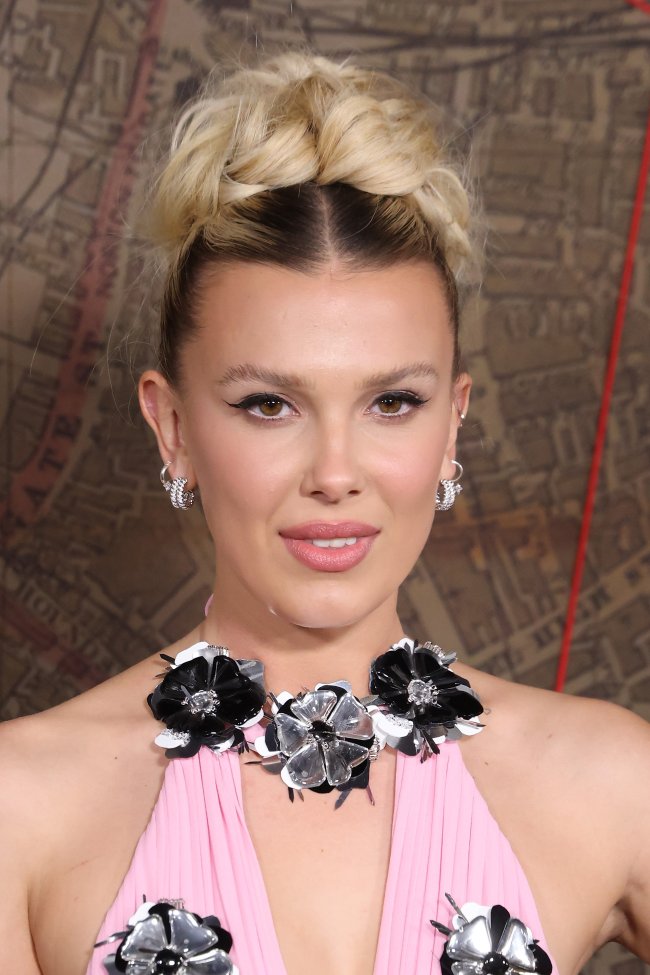 millie-bobby-brown-na-premiere-de-enola-holmes-2 Foto com close no rosto da atriz Millie Bobby Brown na première de seu novo filme 'Enola Holmes 2'. Ela está com um coque fofo, maquiagem simples com delineado e acessórios pratas. Ela olha para frente e sorri levemente.