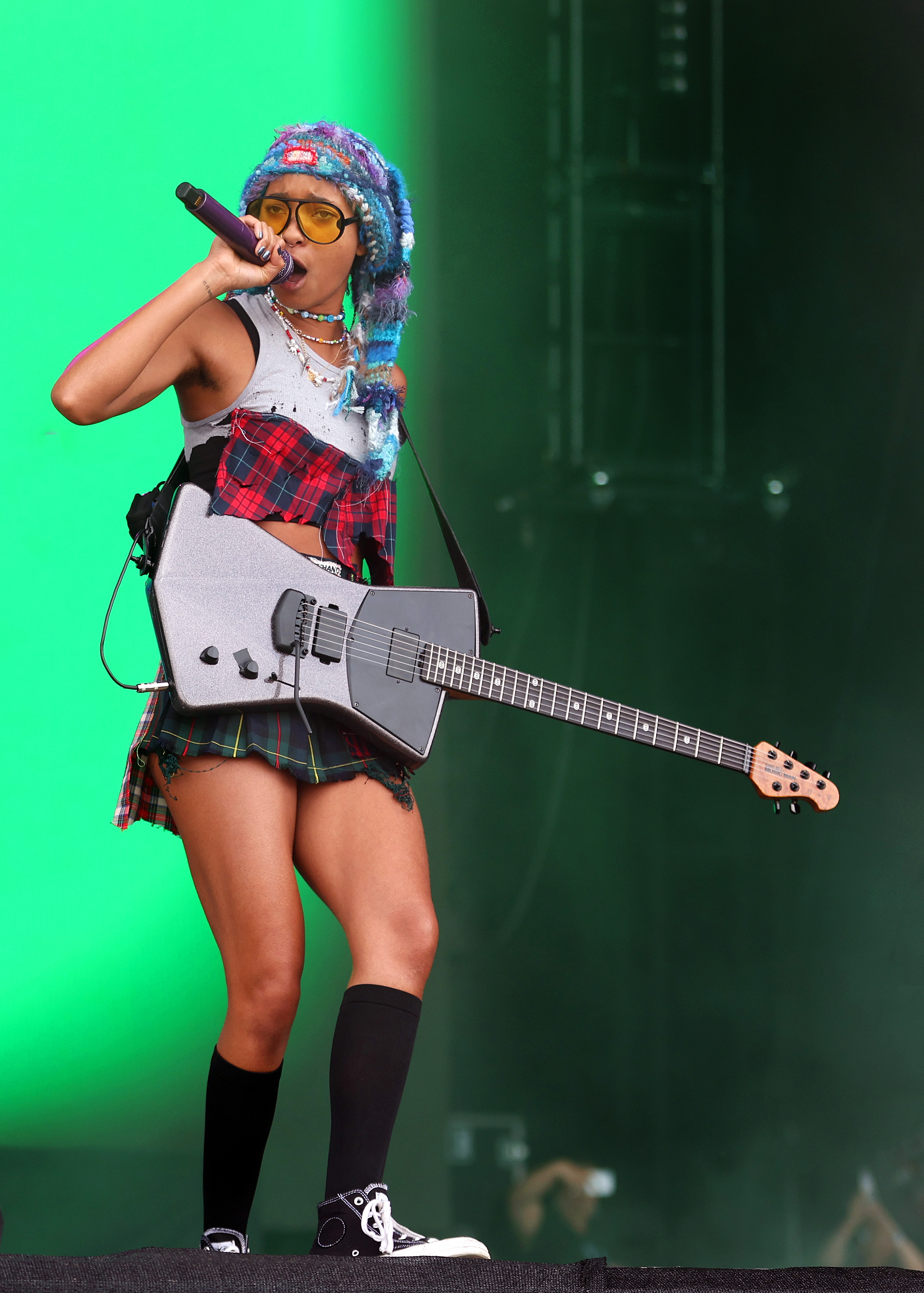 Willow Smith no Reading Festival em agosto de 2022 usando camiseta branca com estampa xadrez, minissaia e meia preta com tênis preto enquanto canta e toca guitarra