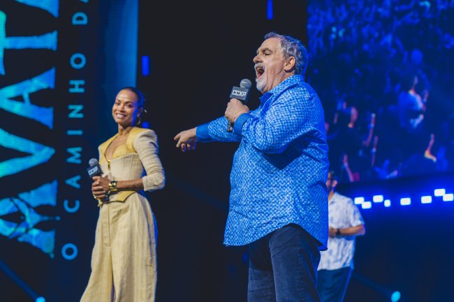 AFCE36F4-7C81-4FA3-B0A5-42CF7FD2A854 Jon Landau sorrindo e apontando para Zoë Saldaña, que sorri enquanto segura microfone no palco da CCXP22
