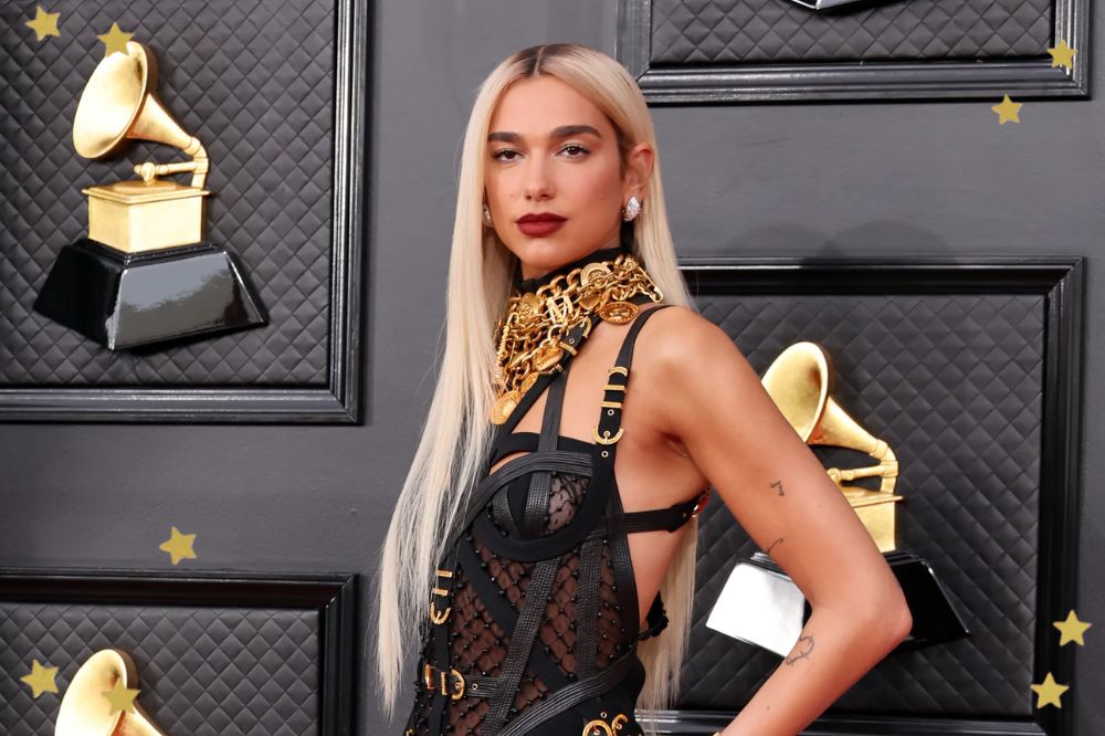 Imagem mostra Dua Lipa no tapete vermelho da 64ª edição do Grammy Awards usando um vestido preto Versace, estilo Bondage, com detalhes dourados. Ela está de lado com a mão esquerda sobre o quadril e olha para a câmera com expressão leve.