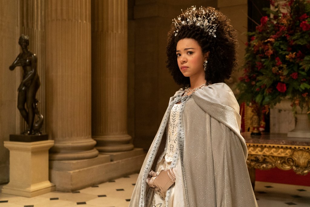 India Ria Amarteifio como a Jovem Rainha Charlotte em cena da série da Netflix; ela está em pé com expressão séria e as mãos na frente do corpo enquanto usa um vestido com uma capa
