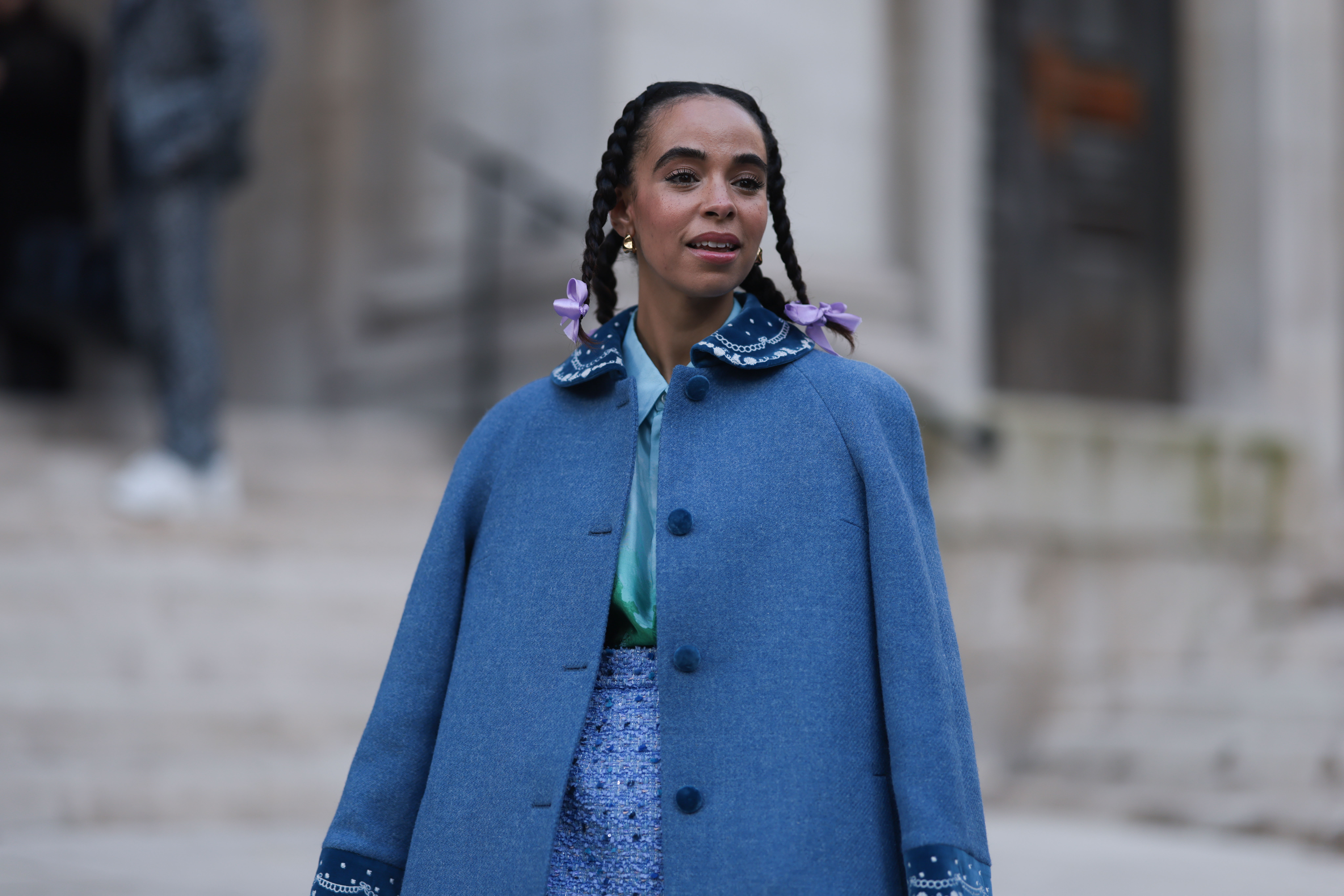 Street Style – LFW February 2023 Mulher usando capa azul e cabelo preso em tranças com laços nas pontas no street style da semana de moda de Londres