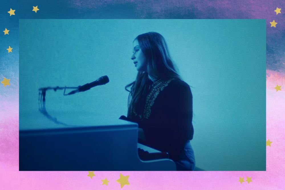 Gabrielle Aplin tocando piano em um ambiente com iluminação azulada; a margem é uma textura nas cores azul, roxo, branco, rosa e lilás com estrelas amarelas decorando a imagem