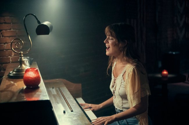 Daisy Jones & The Six Riley Keough como Daisy em Daisy Jones & The Six; ela está tocando piano enquanto canta