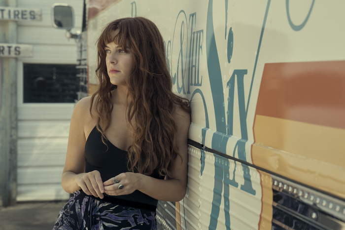 Riley Keough como Daisy em Daisy Jones & The Six; ela está com expressão séria encostada no ônibus de turnê de Daisy Jones & The Six enquanto olha para o lado