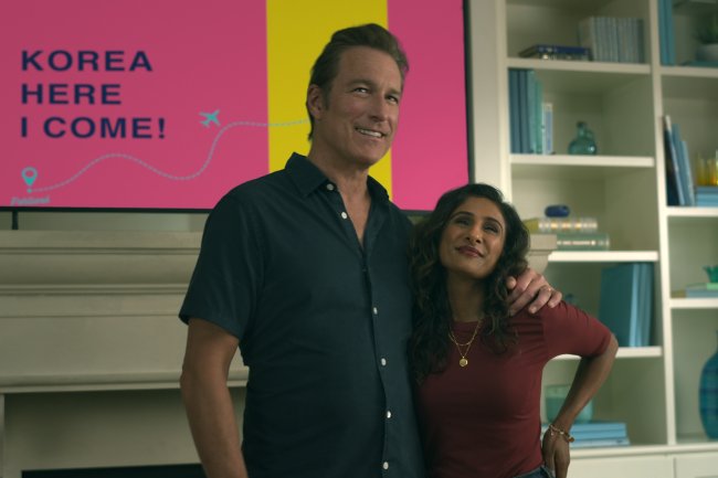 XO,_Kitty_S1_E1_00_05_57_12R John Corbett como Dan Covey e Sarayu Blue como Trina Rothschild em XO, Kitty; eles estão parados e abraçados enquanto sorriem na sala de uma casa