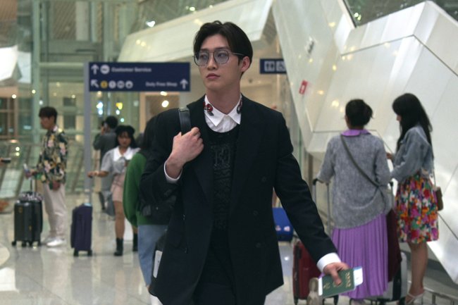 XO,_Kitty_S1_E1_00_06_42_23R Sang Heon Lee como Min Ho em XO, Kitty; ele está com expressão séria andando por um aeroporto com uma mala