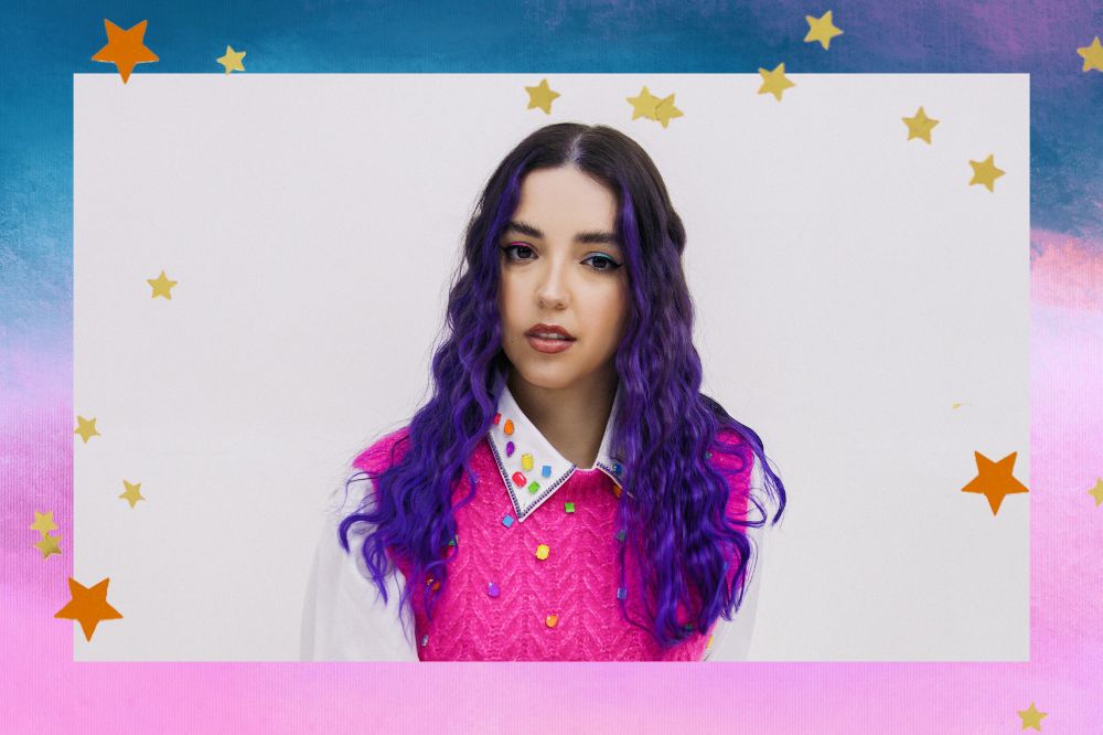 Montagem com fundo azul e rosa. Ana Xisdê está no centro da imagem, ela tem cabelo roxo e veste camisa branca com colete rosa neon. Ana olha diretamente para a camera.