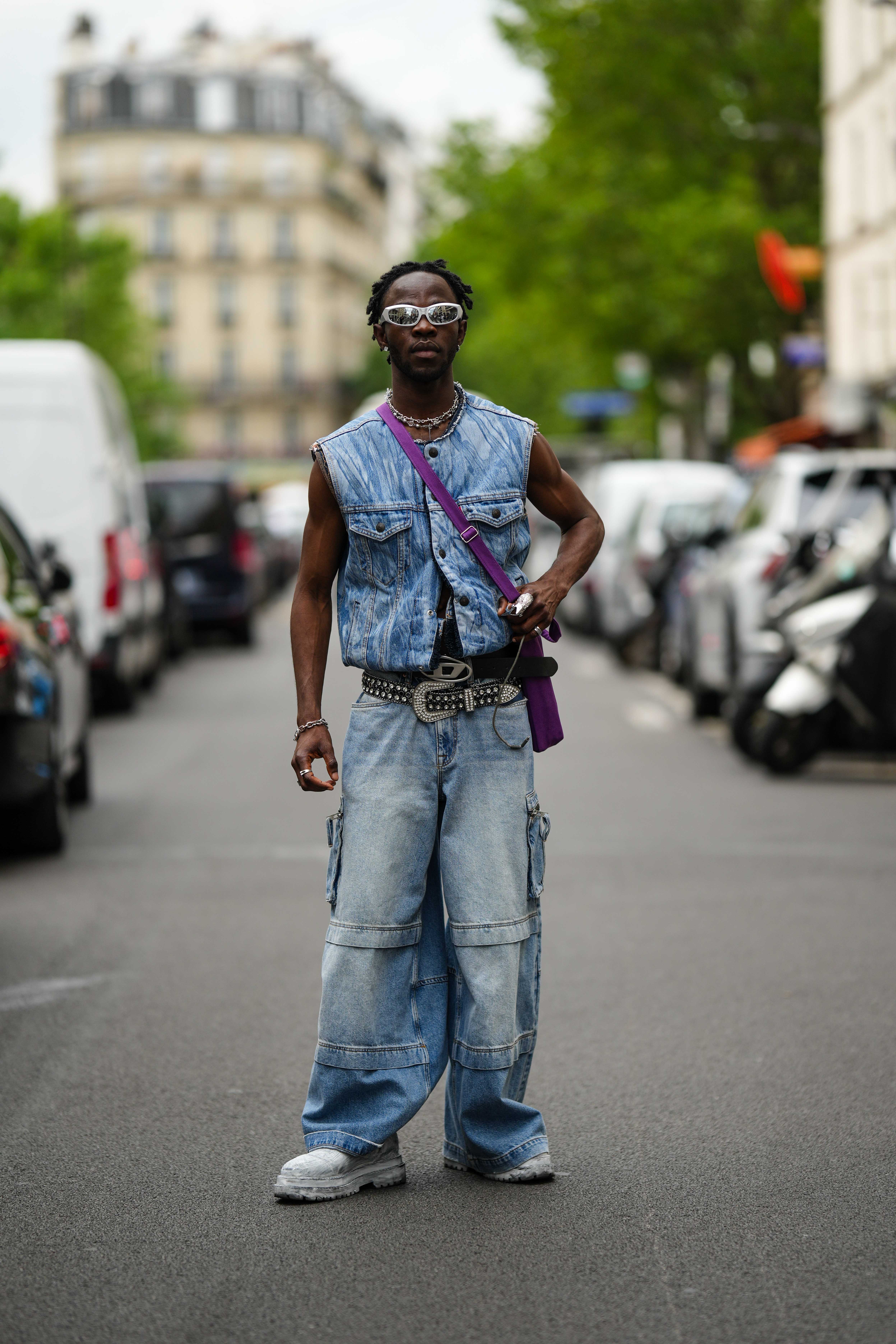 Street Style – Paris Fashion Week – Menswear Spring/Summer 2024 : Day Two Look no street style da semana de moda masculina de primavera-verão 2024 em Paris. Homem usando colete e calça cargo jeans, tênis branco, mix de cintos, bolsa roxa e óculos metalizados