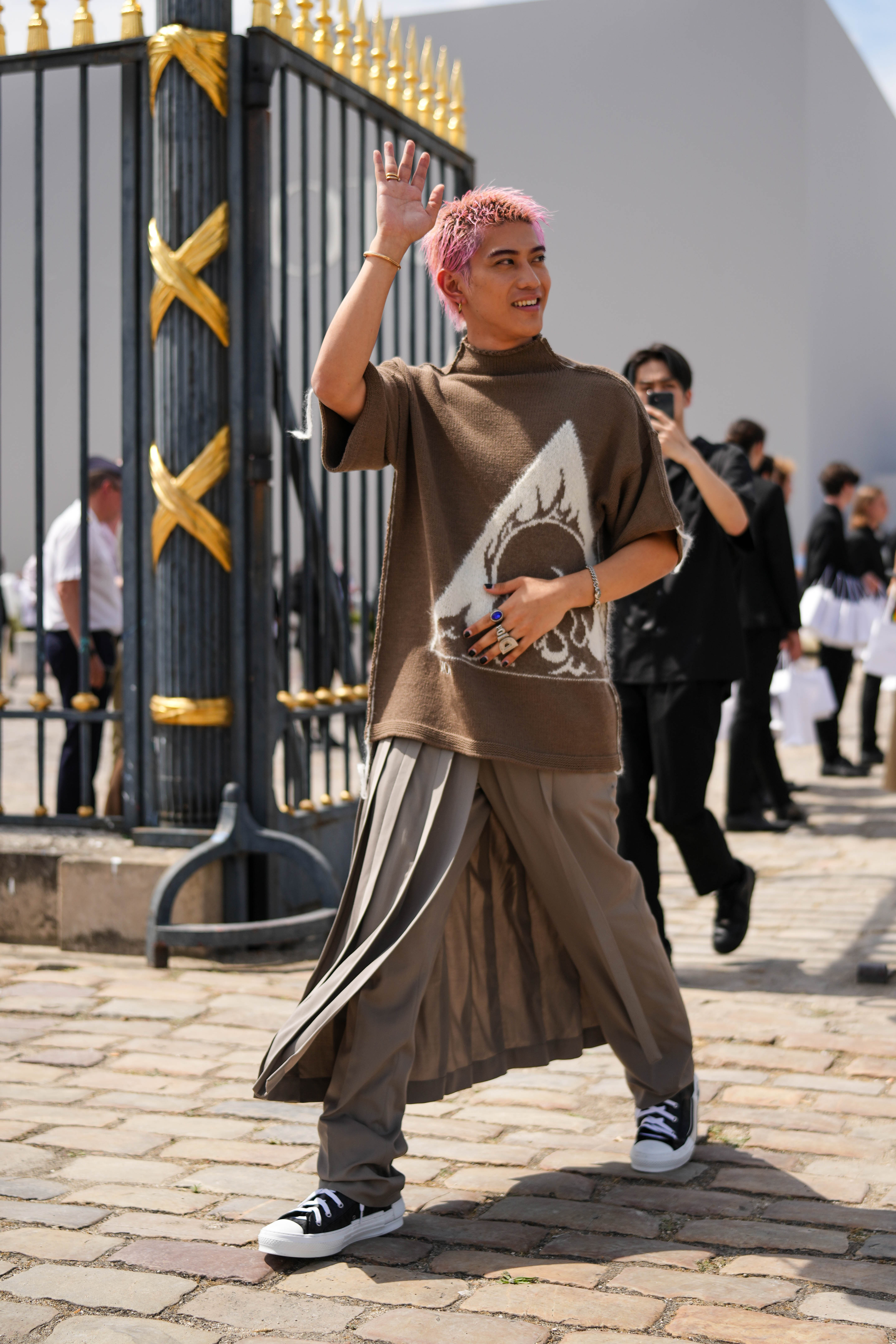 Street Style – Paris Fashion Week – Menswear Spring/Summer 2024 : Day Four Look do Yamato no street style da semana de moda masculina de primavera-verão 2024 em Paris. Ele usa blusa de tricô marrom, calça em tom parecido com saia plissada por cima e tênis