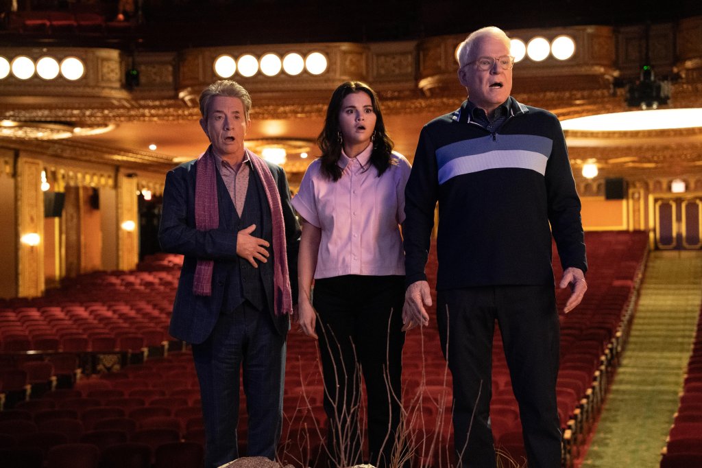 Cena da 3ª temporada de Only Murders in The Bulding com Oliver, Mabel e Charles, interpretados por Martin Short, Selena Gomez e Steve Martin, respectivamente, com expressões de surpresa e choque enquanto estão em um palco de teatro com as cadeiras do público ao fundo