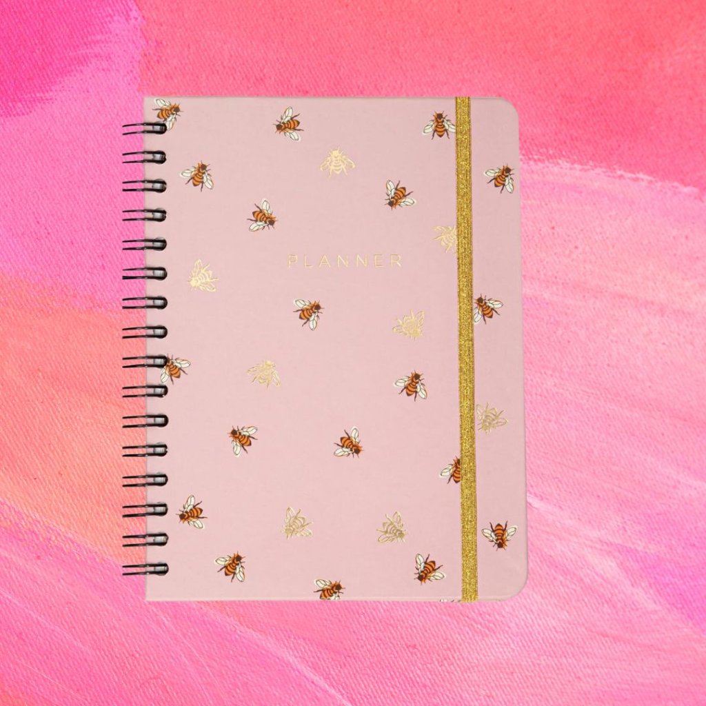 planner Planner rosa com detalhes de abelhas na estampa e uma tira dourada; o fundo é uma textura em tons de rosa e branco