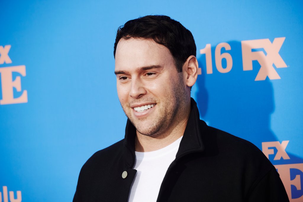 Scooter Braun posando para foto e sorrindo com um backdrop azul ao fundo