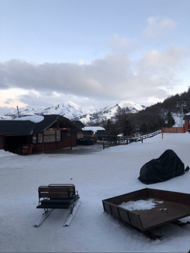 A imagem mostra a neve cobrindo o chão. Na frente aparece um trenó e no fundo montanhas com seus topos cobertos de neve