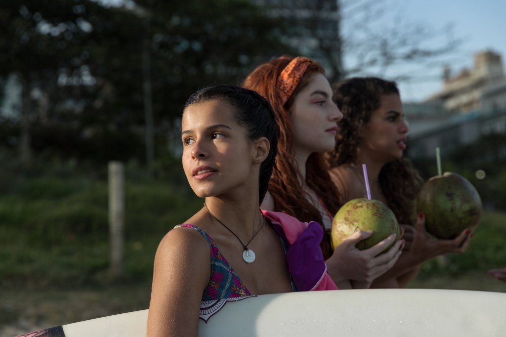 Atrizes em cena de Tudo Igual... SQN; elas estão na praia com a protagonista fotografada de perfil olhando para trás por cima do ombro e sorrindo levemente