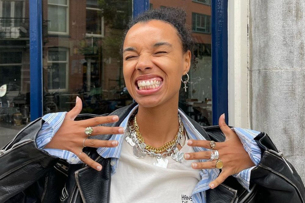 Menina com jaqueta preta, camisa azul e camiseta branca. Ela está sorrindo e com as mão abertas. Usa muitos acessórios. prata e dourado