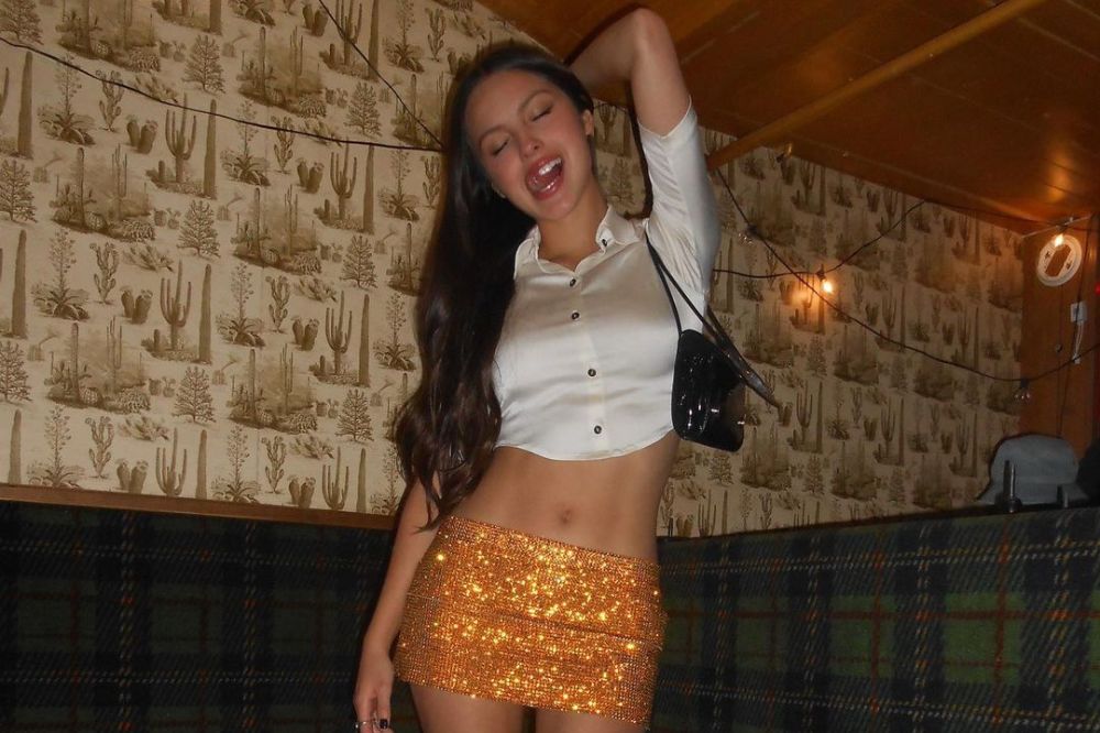 Foto da Olivia Rodrigo mostrando a língua com cabelo solto, saia laranja e camisa branca