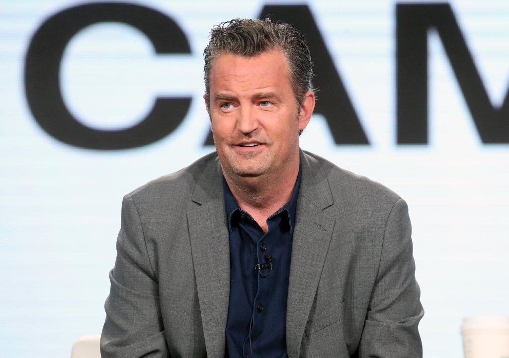 Matthew Perry está usando um terno cinza escura e camisa azul escura