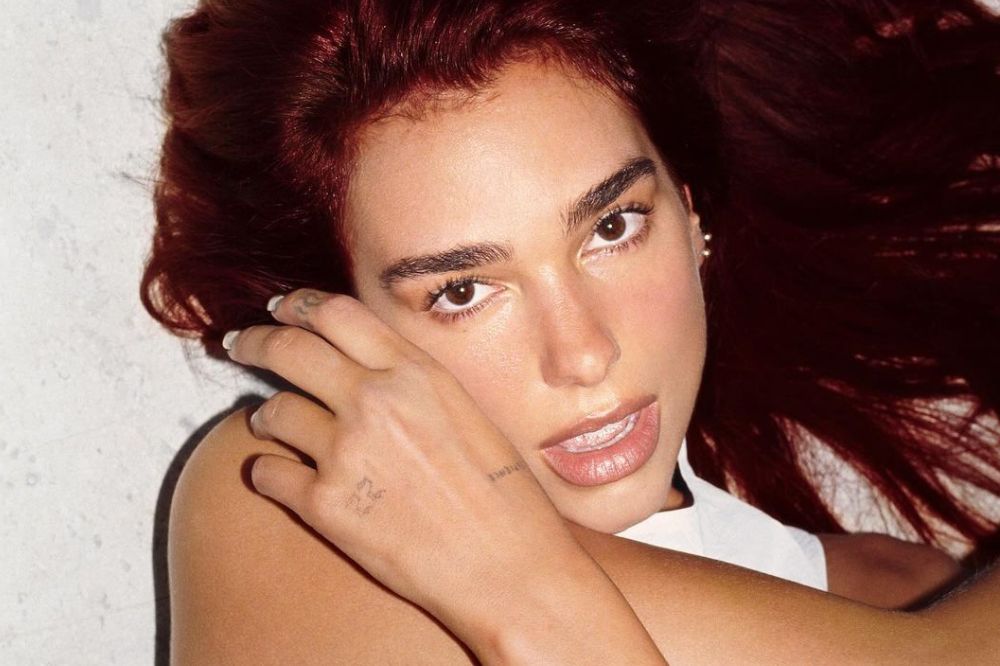 Dua Lipa posando para foto com cabelo ruivo; ela está com os braços entrelaçados na frente do corpo e uma das mãos próxima ao rosto