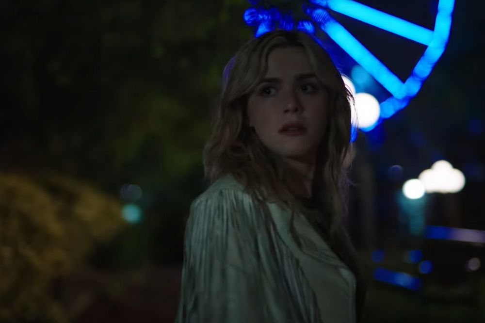 Kiernan Shipka em cena de Dezesseis Facadas; ela está olhando para trás por cima do ombro durante a noite com uma expressão assustada e atenta; ao fundo, está uma roda-gigante com luzes azuis e árvores